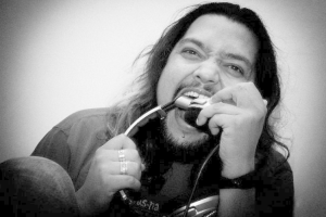 Ein Mann mit langen Haaren und Bart singt in ein Mikrofon vor einer Wand, trägt ein schwarzes T-Shirt und Jeans und hat einen ernsten Gesichtsausdruck.