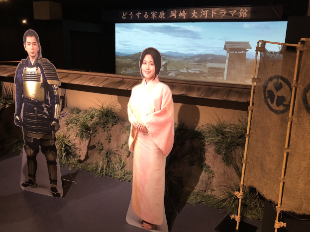 Eine Frau in einem rosafarbenen Kimono und ein Mann in einem Samurai-Kostüm, die gemeinsam in einer Szene eines japanischen Films lächeln, mit Pflanzen, einem Bildschirm und Lichtern im Hintergrund.