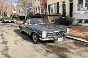 Ein 1971 Mercedes-Benz 280SL, das auf einer Stra├če in einer Stadt geparkt ist, umgeben von Geb├Ąuden, Pflanzen und urbanen Elementen.