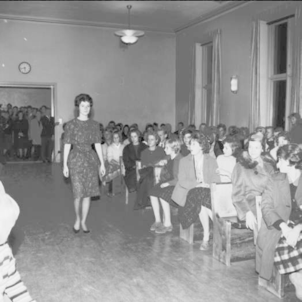Eine Frau in einem schwarzen und weißen Kleid läuft über einen Laufsteg während einer Modenschau für Frauenwahlrecht, mit einer Menge, die auf Stühlen sitzt und einer Uhr und Lampen an der Wand.