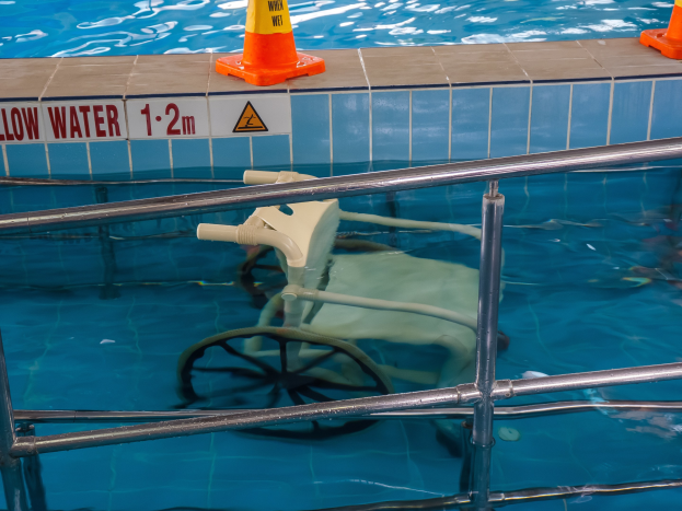 Schwimmbad mit einem Stuhl in der Mitte, umgeben von Sicherheitskegeln und einem Geländer, mit einem Schild an der Wand, das verfügbare Schwimmausrüstung anzeigt.