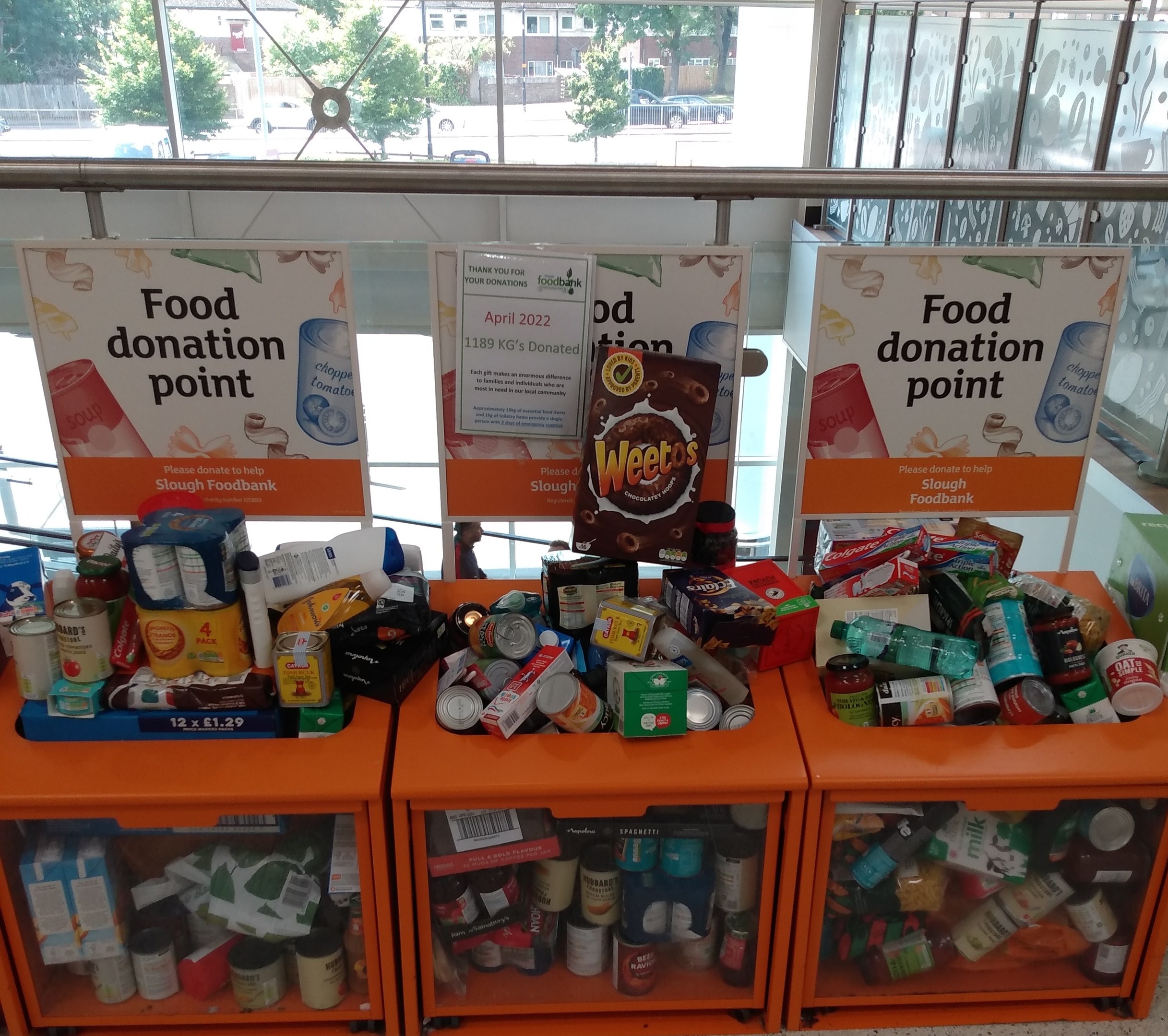 Orangene Schübe mit Lebensmittelspenden, darunter Flaschen, Dosen und Kisten, mit Informationsschildern und Blick auf Fahrzeuge, Bäume, Gebäude und Himmel durch Glasfenster.