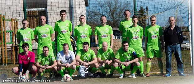 Eine Gruppe von Männern in grünen T-Shirts steht auf einem Fußballfeld mit einem Ball und einem Tor, einige in der Hocke, mit Bäumen, Gebäuden und Fahrzeugen im Hintergrund.