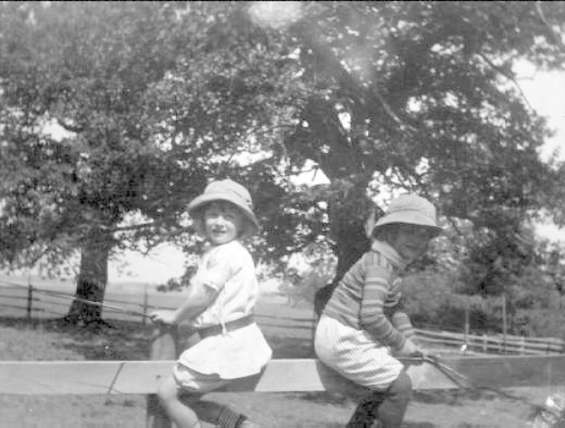 Zwei junge Jungs in Hüten und Baseball-Uniformen stehen in einem grünen Park und halten Schläger, mit Bäumen und einem Zaun im Hintergrund, in Schwarz-Weiß.