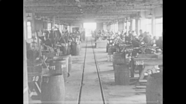 Schwarzes und weißes Foto einer Fabrikhalle während der industriellen Revolution, das Arbeiter bei der Bedienung von Maschinen zeigt, nummerierte Fässer, eine Eisenbahnschiene, tragenden Säulen und Deckenbeleuchtung.