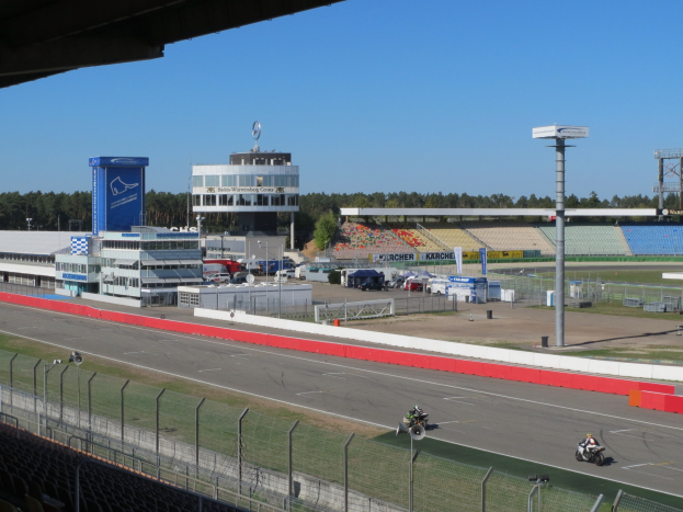 Motorradfahrer auf einer Rennstrecke mit Absperrung, Fahrzeugen, Gebäude, Beschilderung, Stadion, Beleuchtung, Bäumen und bewölktem Himmel.
