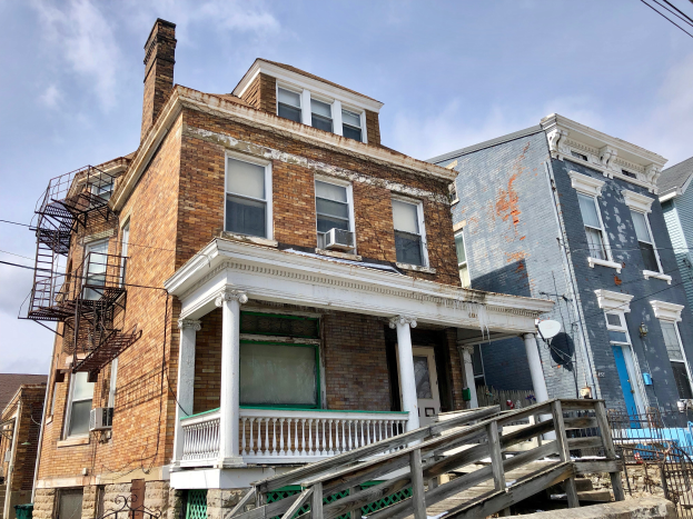 Eine Reihe von baufälligen Häusern an einer Straßenecke, mit zerbrochenen Fenstern, bröckelnden Wänden, Geländern, Stufen, einem Zaun, einer Satellitenschüssel, Drähten und einem bewölkten Himmel.