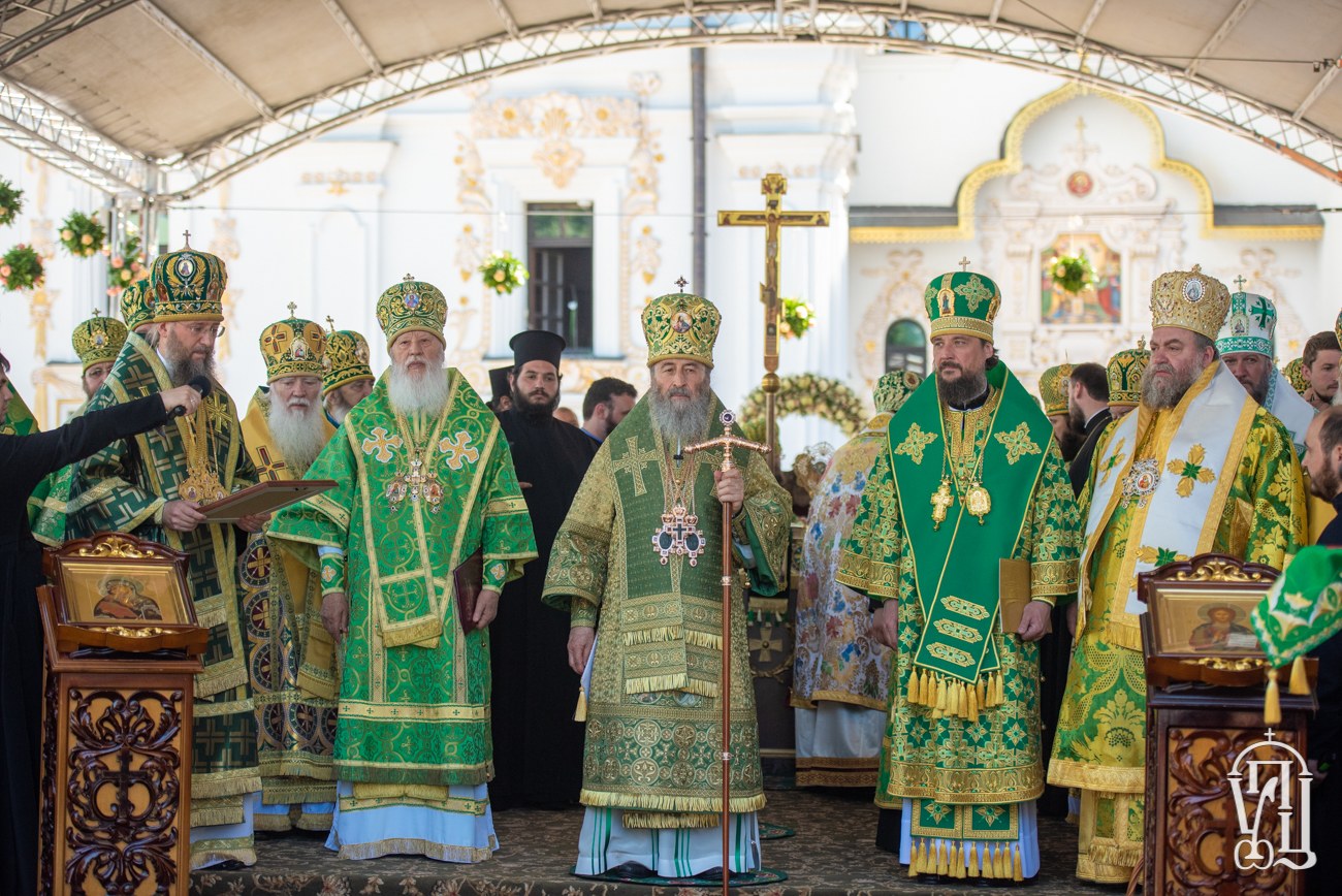 Gruppe von Priestern vor einer Kirche während einer religiösen Zeremonie, einer Person mit einem Buch und einem Mikrofon auf der linken Seite des Bildes, einem Kreuzsymbol, Blumen und einem Gebäude im Hintergrund.