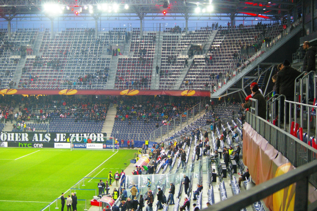Großes Stadion voller Zuschauer bei einem Fußballspiel mit Geländer, Banner und leerem Sitzbereich im Hintergrund.