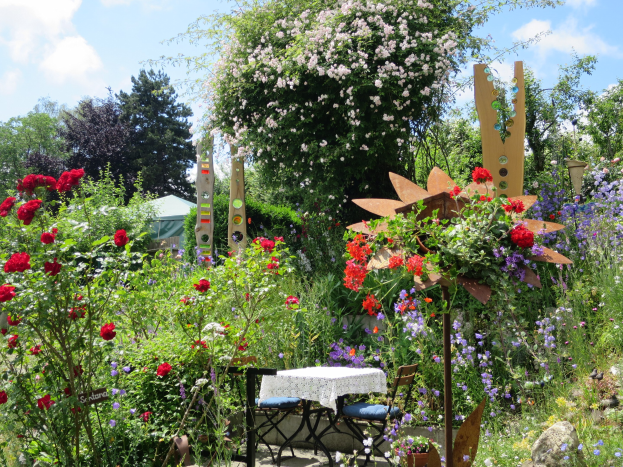 Ein Garten auf dem Chelsea Flower Show mit bunten Blumen und Pflanzen, einem Tisch mit einer Decke und Stühlen in der Mitte, Bäumen und einem bewölkten Himmel im Hintergrund.