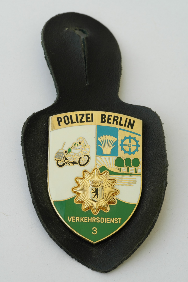 Ein schildförmiges Polizeabzeichen mit 'Polizei Berlin' in fetter schwarzer Schrift, das ein weißes Stern auf blauem Hintergrund und einen weißen Rand aufweist und auf einer Oberfläche liegt.