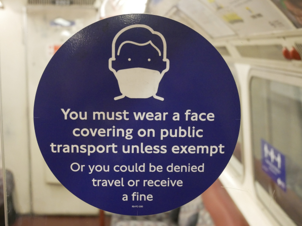 Schriftzug an einer Zugtür, der das Tragen von Mund-Nasen-Schutz auf öffentlichen Verkehrsmitteln mit Strafen für Nichtbefolgung angibt, mittig in einem Glasfenster.