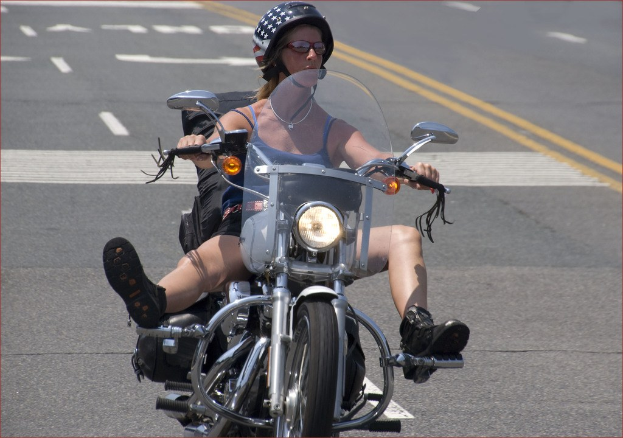 Professionelle Radsportlerin Katie Archibald mit Helm, Schutzbrille, Handschuhen und Schuhen fährt auf einem Fahrrad eine Straße entlang neben einer Menge von Menschen, mit einem Banner, einer Wand, Bäumen und einem Gebäude im Hintergrund.
