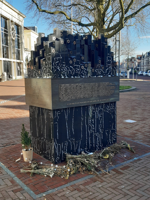 Denkmal für Holocaust-Opfer in einem Stadtplatz mit einem Baum vorne, einem Topfgewächse auf dem Boden, verstreuten trockenen Blättern und Gebäuden, Fahrzeugen, Masten und Bäumen im Hintergrund.