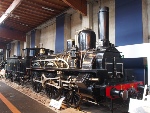 Alte Dampflokomotive in einem Museums Setting mit Schienen, Informationsschildern und anderen Ausstellungsstücken im Hintergrund.