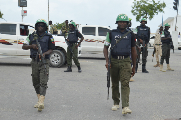 Eine Gruppe von Polizisten in Helmen, die Waffen halten, geht eine Straße entlang mit Fahrzeugen, Laternenmasten, Verkehrsampeln, Texttafeln, Bäumen, Gebäuden und einem bewölkten Himmel im Hintergrund.
