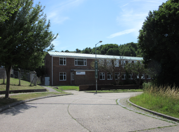 Ein Schulgebäude mit Fenstern, einem Straßenschild, einem Schild, einer Baumgruppe, Gras, einem Zaun und einem bewölkten Himmel, das sich in einem Park mit saftigem Grün befindet.