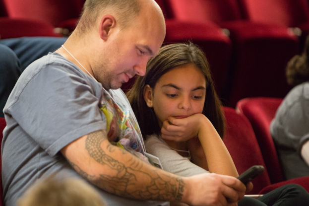 Ein Mann und ein junges Mädchen sitzen in einem Kino, beide schauen auf ein Handy, wobei der Mann es hält und das Mädchen es genau betrachtet.