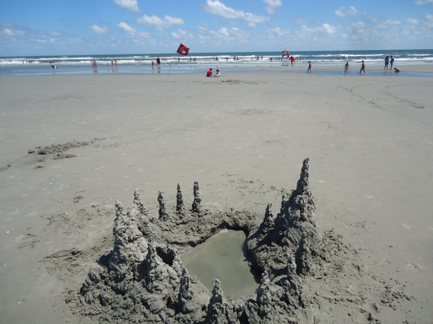 Ein Sandburgenbau am Strand mit Menschen, Wasser und einer Fahne im Hintergrund unter einem bewölkten Himmel.