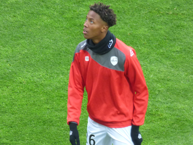 Ein junger Mann in einer roten Jacke und weißen Shorts steht auf einem Fußballfeld, trägt Baseball-Handschuhe und schaut entschlossen nach rechts.