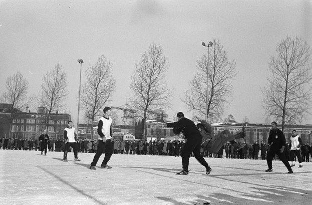 Eine Gruppe von Menschen, die im Schnee Eishockey spielen, mit Bäumen, Gebäuden, Laternenmasten und einem klaren Himmel im Hintergrund, dargestellt in Schwarz-Weiß.