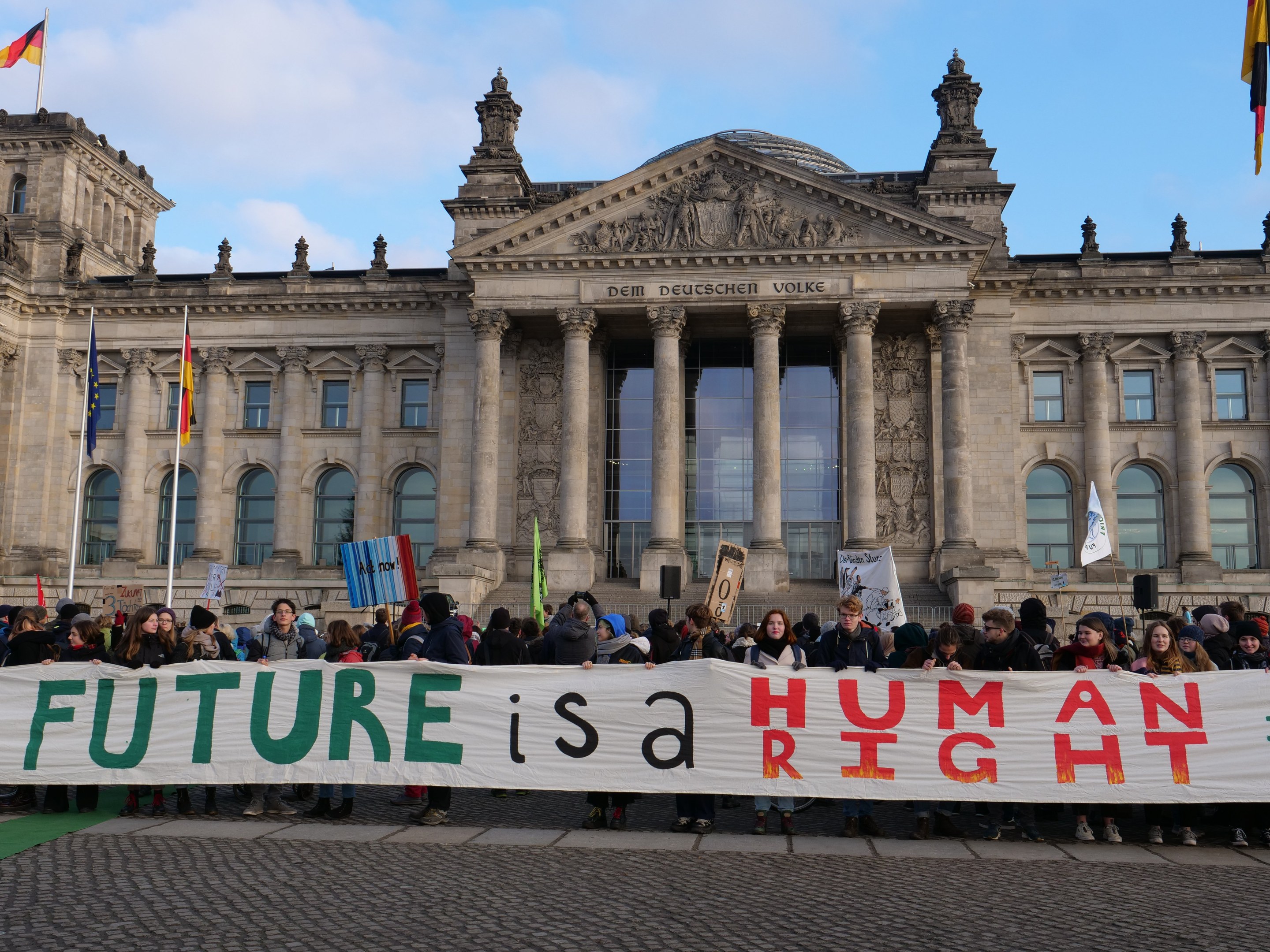 Gruppe von Menschen mit Transparent 'Zukunft ist ein Menschenrecht' vor dem Reichstagsgebäude in Berlin, Deutschland, mit bewölktem Himmel und umliegenden Fahnenmasten.