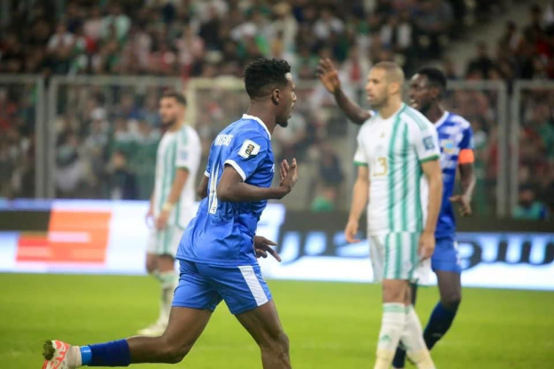 Ein Fußballer in einem blauen und weißen Trikot feiert mit erhobenen Armen, umgeben von einer Gruppe in weißen und grünen Trikots, mit einem unscharfen Hintergrund, der ein weißes und blaues Brett und ein Geländer zeigt.