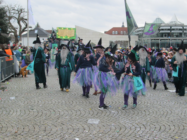 Gruppe von Menschen in Hexenkostümen, die eine Kopfsteinpflasterstraße entlanggehen, tragen schwarze und lila Kleider und Hüte, einige halten Fahnen, mit einem Geländer auf der linken Seite und Gebäuden, Bäumen und einem klaren blauen Himmel im Hintergrund.