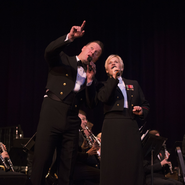 Ein Mann und eine Frau in Fracks, die Mikrofone auf einer Bühne halten, mit einer Sitzreihe von Musikern dahinter in einer dunklen Umgebung.
