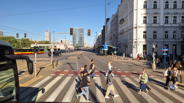 Eine Gruppe von Menschen, die eine Straße an einer Ampel überqueren, mit einem Bus auf der linken Seite, Fahrzeugen auf der Straße, Verkehrszeichen, Laternenmasten, Gebäuden, Bäumen und einem klaren blauen Himmel.