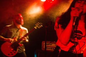 Eine Frau singt in ein Mikrofon auf der Bühne, wöhrend ein Mann eine Akustikgitarre im Hintergrund spielt, mit Deckenlampen, die die lebendige Szenerie beleuchten.