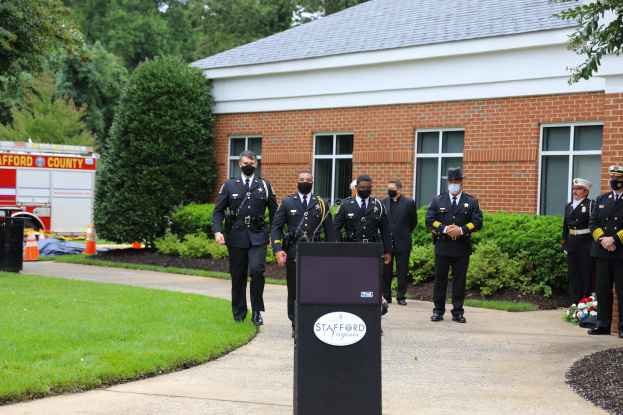 Eine Gruppe von Polizisten in Uniform und Masken steht vor einem Gebäude mit Fenstern und Türen, mit einem Podium, Mikrofon und Texttafel im Vordergrund und einem Löschfahrzeug, Bäumen, Pflanzen, Blumen und anderen Gegenständen im Hintergrund.