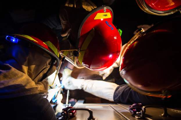 Feuerwehrleute in Helmen und Handschuhen an einem Feuerwehrauto in einer dunklen Umgebung, mit einem Tisch voller Gegenstände auf der rechten Seite.
