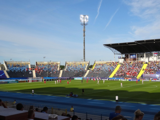 Ein Fußballspiel im Gange auf einem großen Stadionfeld mit Zuschauern auf den Rängen, beleuchtet von Flutlicht unter einem sichtbaren Himmel.