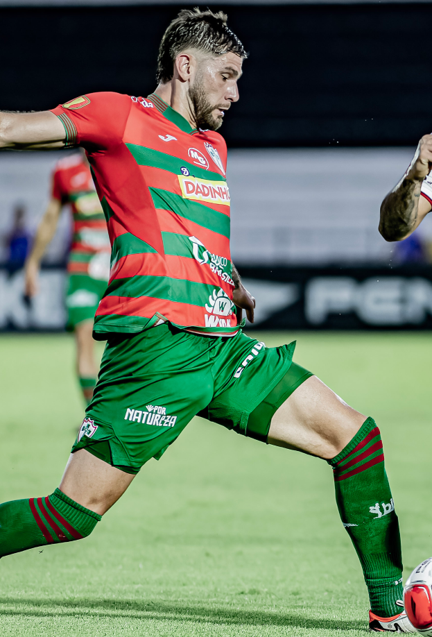 Ein Mann in roter und grüner Uniform kickt einen Fussball auf einem Rasenfeld, mit einer anderen Person im Hintergrund und einer Tafel, die "Santos vs Vitoria Guimaraes" anzeigt.
