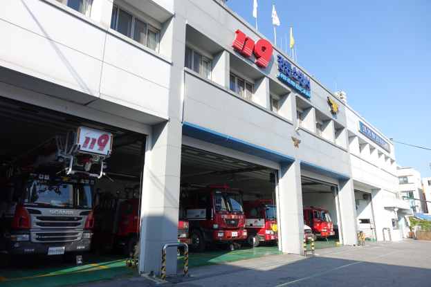 Feuerwache mit mehreren Feuerwehrautos vor der Tür, mit Fenstern, Säulen und Namensschildern, Flaggen im Vordergrund und anderen Gebäuden gegen einen klaren blauen Himmel.