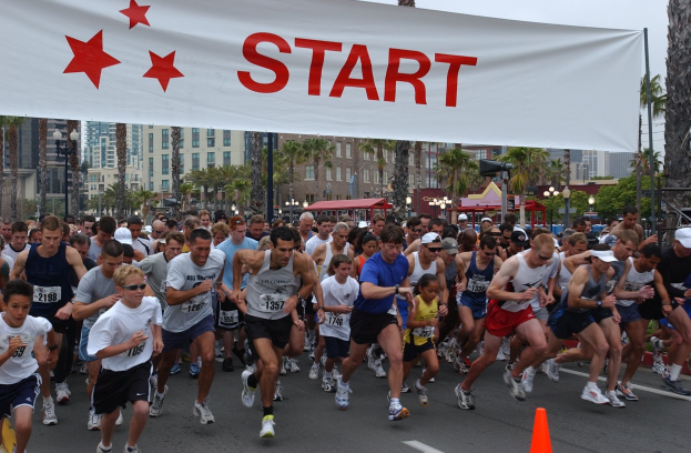 Eine Gruppe von Läufern bei einem Marathon mit einem Verkehrskegel im Vordergrund und einem Textbanner im Hintergrund, vor Bäumen, Laternenmasten, Gebäuden und einem klaren blauen Himmel.