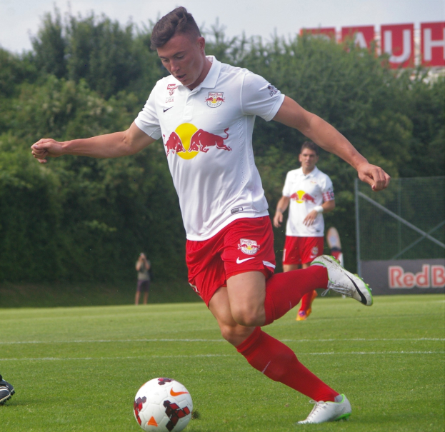 Ein Mann in einem Fußballtrikot schießt einen Ball auf einem Rasenfeld mit Bäumen und einem klaren blauen Himmel im Hintergrund, wobei ein "RB Leipzig"-Schild sichtbar ist.