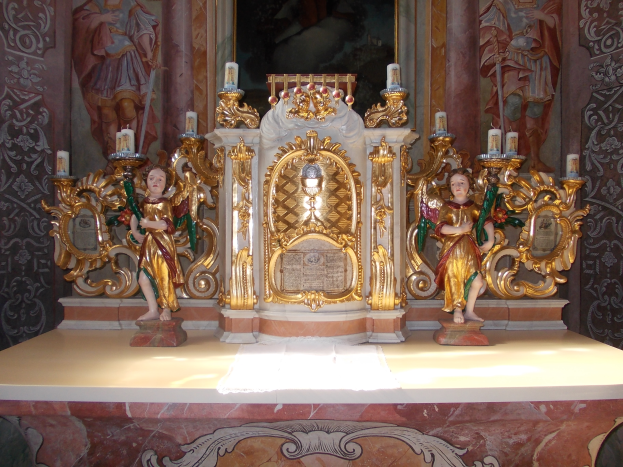 Ein Altar in einer Kirche mit Skulpturen von Menschen und Kerzen auf Ständen, mit einem Gemälde an der Wand dahinter, das eine feierliche Atmosphäre schafft.