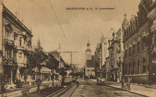 Ein Schwarz-Weiß-Foto einer Stadtstraße in Brandenburg, Deutschland, mit Gebäuden, Bäumen, Polen, Drähten, Menschen, Karren und Bahnschienen, sowie Text oben und einem sichtbaren Himmel im Hintergrund.