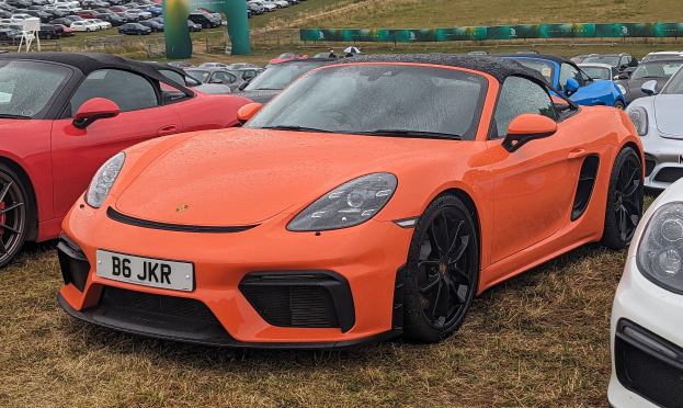 Roter Porsche 718 Boxster GTS auf der Goodwood Festival of Speed, zusammen mit anderen Autos, einem Banner, Bäumen und einem klaren blauen Himmel im Hintergrund.
