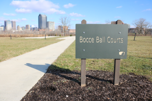 Platte mit Text "Bocce Ball Courts in Indianapolis, Indiana" auf Gras neben einem Weg, Bäumen, Pfählen, einem Zaun, Gebäuden und einem bewölkten Himmel.