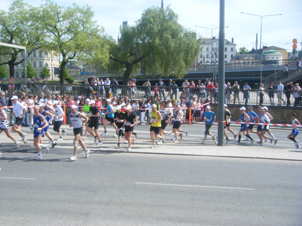 Gruppe von Läufern beim Marathon auf einer Straße mit einem Zielpfosten und einer Ziellinie, umgeben von einer Metallabsperrung und Zuschauern sowie städtischen Elementen wie Pfosten, Schildern, einer Brücke, Gebäuden, Bäumen und einer bewölkten Himmel.