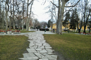 Ein Park mit einem gewundenen Steinweg, grünem Gras, Bäumen, Bänken, Tischen, Gebäuden und einem klaren blauen Himmel.