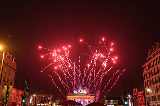 Eine nächtliche Straßenansicht in Berlin am Silvesterabend, mit Gebäuden, Bäumen, Laternen, Ampeln, Schildern, Zelten, Menschen und einem Feuerwerk am Himmel.
