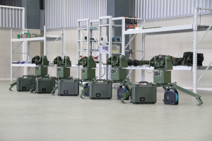 Eine Gruppe von Militärrobotern, die auf einem Boden mit Koffern und anderen Gegenständen auf umliegenden Gestellen positioniert sind, vor einer Wand mit einem Geländer im Hintergrund.