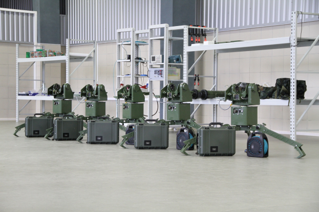 Eine Gruppe von Militärrobotern, die auf einem Boden mit Koffern und anderen Gegenständen auf umliegenden Gestellen positioniert sind, vor einer Wand mit einem Geländer im Hintergrund.