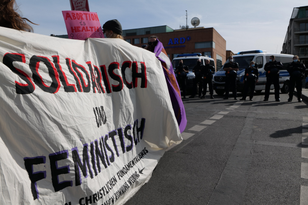 Eine Gruppe von Menschen marschiert die Straße entlang, hölt eine 'Solidarität und Feminismus'-Fahne, mit geparkten Fahrzeugen und Gebäuden im Hintergrund unter einem klaren blauen Himmel.