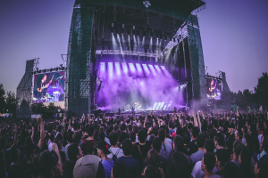 Eine große Menschenmenge bei einem Musikfestival schaut auf einer hell beleuchteten Bühne mit Instrumenten und Bildschirmen, die vor einem Gebäude, Bäumen und einem klaren blauen Himmel aufgebaut sind.