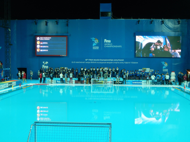 Gruppe von Menschen, die um ein Hallen-Schwimmbecken mit Netz am Boden stehen, mit einer Wand im Hintergrund, die den Text und Logos 'FINA World Championships 2015 Kazan' anzeigt, sowie Trägern und Lampen.
